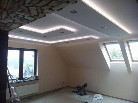 Nowoczesny pokój na poddaszu z oknami dachowymi i dekoracyjnym sufitem podwieszanym z oświetleniem LED, fragment ściany wykończony kamieniem dekoracyjnym, widoczna piła ukośnica.