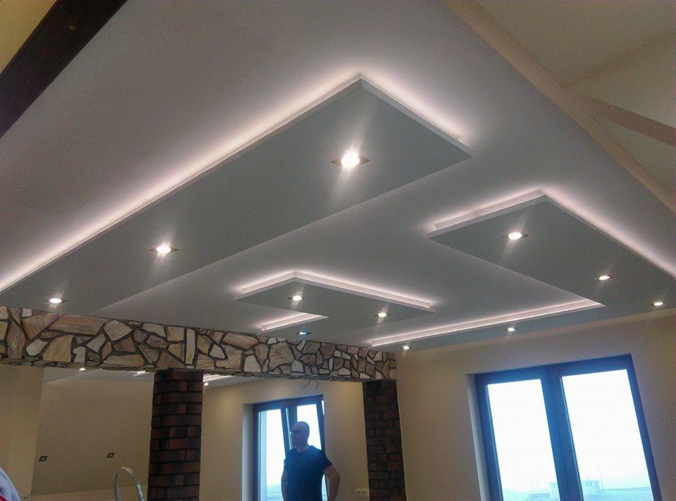Wielopoziomowy sufit podwieszany z oświetleniem LED w kształcie prostokątów i kwadratów, z widocznymi fragmentami ściany wykończonej kamieniem dekoracyjnym i ceglanymi filarami.