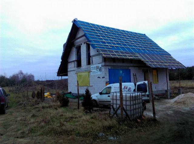 Niedokończony dom jednorodzinny z niebieską folią na dachu i częściowo zamurowanymi oknami, widoczny samochód dostawczy z logo firmy budowlanej.