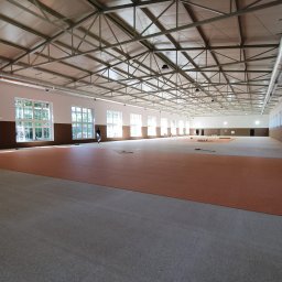 JPB sp. z o.o. - Przestronna hala w trakcie wykańczania, z widoczną stalową konstrukcją dachu, rzędami okien w białych ramach i częściowo ułożoną, pomarańczową wykładziną podłogową.