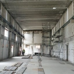 JPB sp. z o.o. - Wnętrze hali w trakcie budowy z widocznymi rusztowaniami, płytami na podłodze i pracownikiem w oddali.