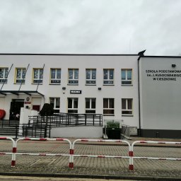 JPB sp. z o.o. - Dwukondygnacyjny budynek Szkoły Podstawowej im. J. Kusocińskiego w Cieszkowie z płaskim dachem, białymi ścianami i żółtymi obramowaniami okien, ozdobiony świątecznymi dekoracjami w oknach, widoczny...