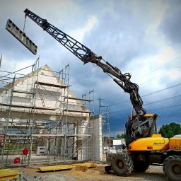 JPB sp. z o.o. - Żółta maszyna budowlana Mecalac podnosi element szalunkowy na budowę domu z białego bloczka, widoczne rusztowania i pochmurne niebo w tle.