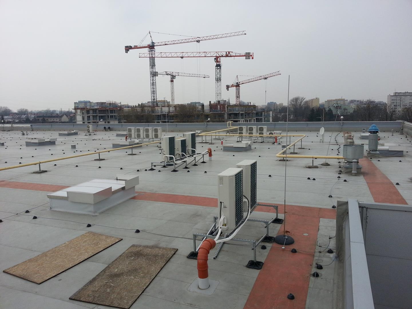 Widok na płaski dach budynku z instalacjami wentylacyjnymi, rurami, świetlikami i elementami konstrukcyjnymi, na tle budowy z żurawiami.