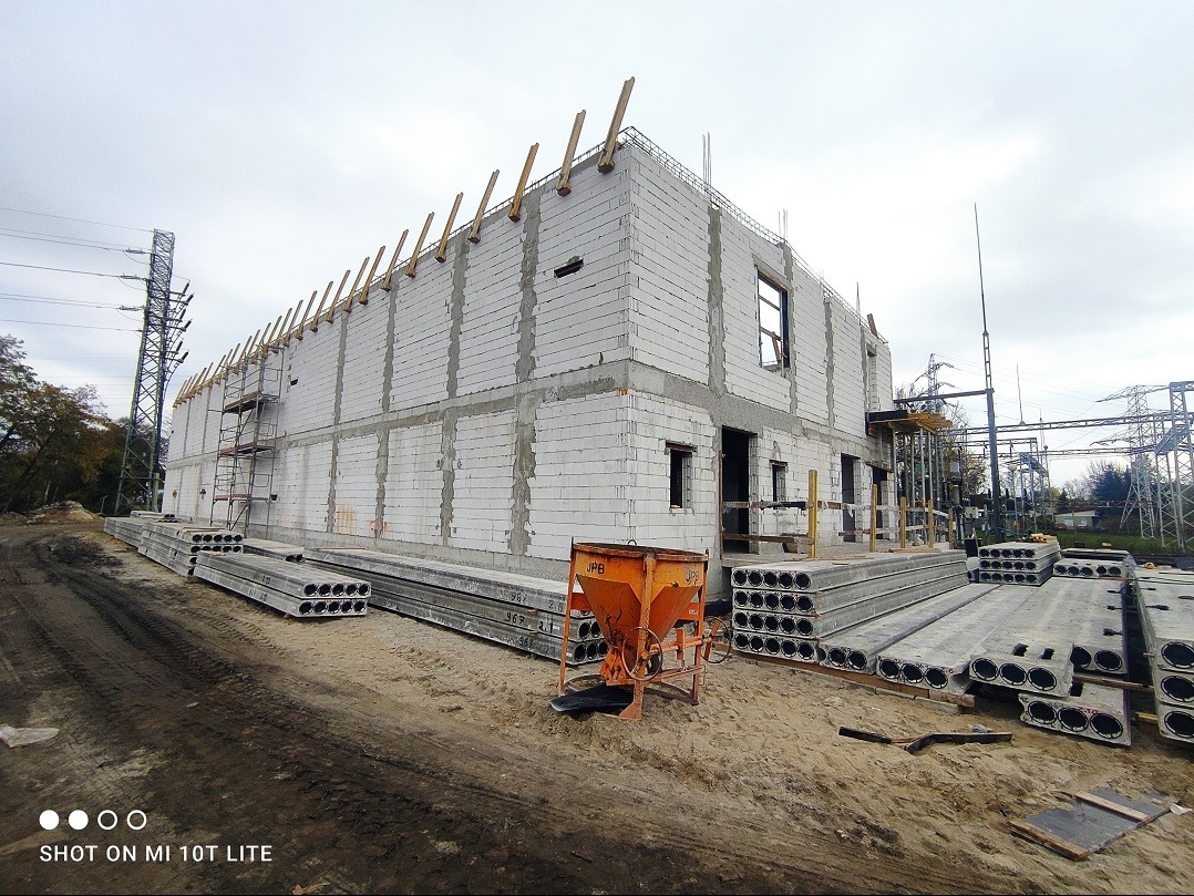 Dwukondygnacyjny budynek w stanie surowym, wznoszony z białych bloczków, z widocznymi drewnianymi elementami konstrukcyjnymi oraz stertami prefabrykowanych płyt stropowych na placu budowy; w tle...