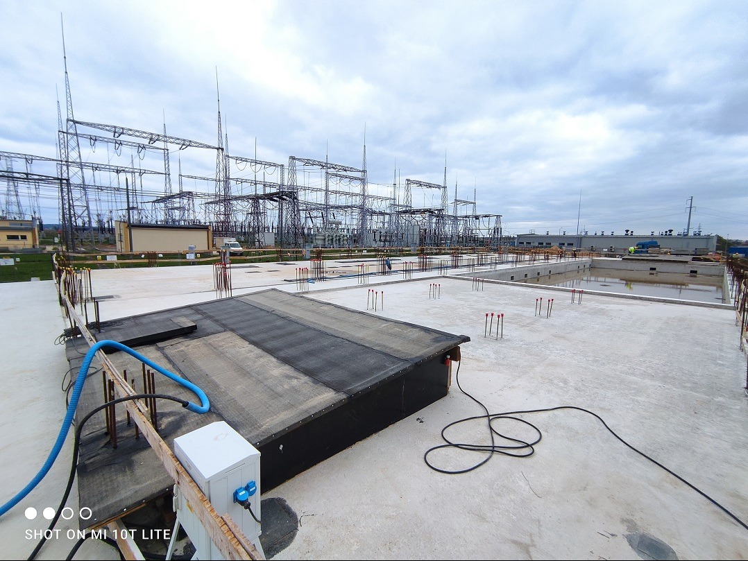 Betonowa płyta fundamentowa pod budynek przemysłowy z wystającymi prętami zbrojeniowymi, widoczne elementy instalacji i w tle słupy wysokiego napięcia.