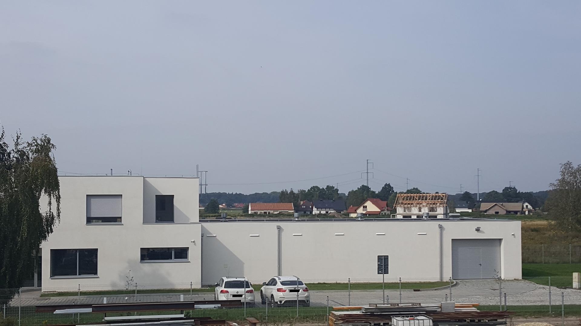 Nowoczesny, biały budynek hali produkcyjnej z dwiema zaparkowanymi, białymi osobówkami na tle budowy domu jednorodzinnego w stanie surowym otwartym.