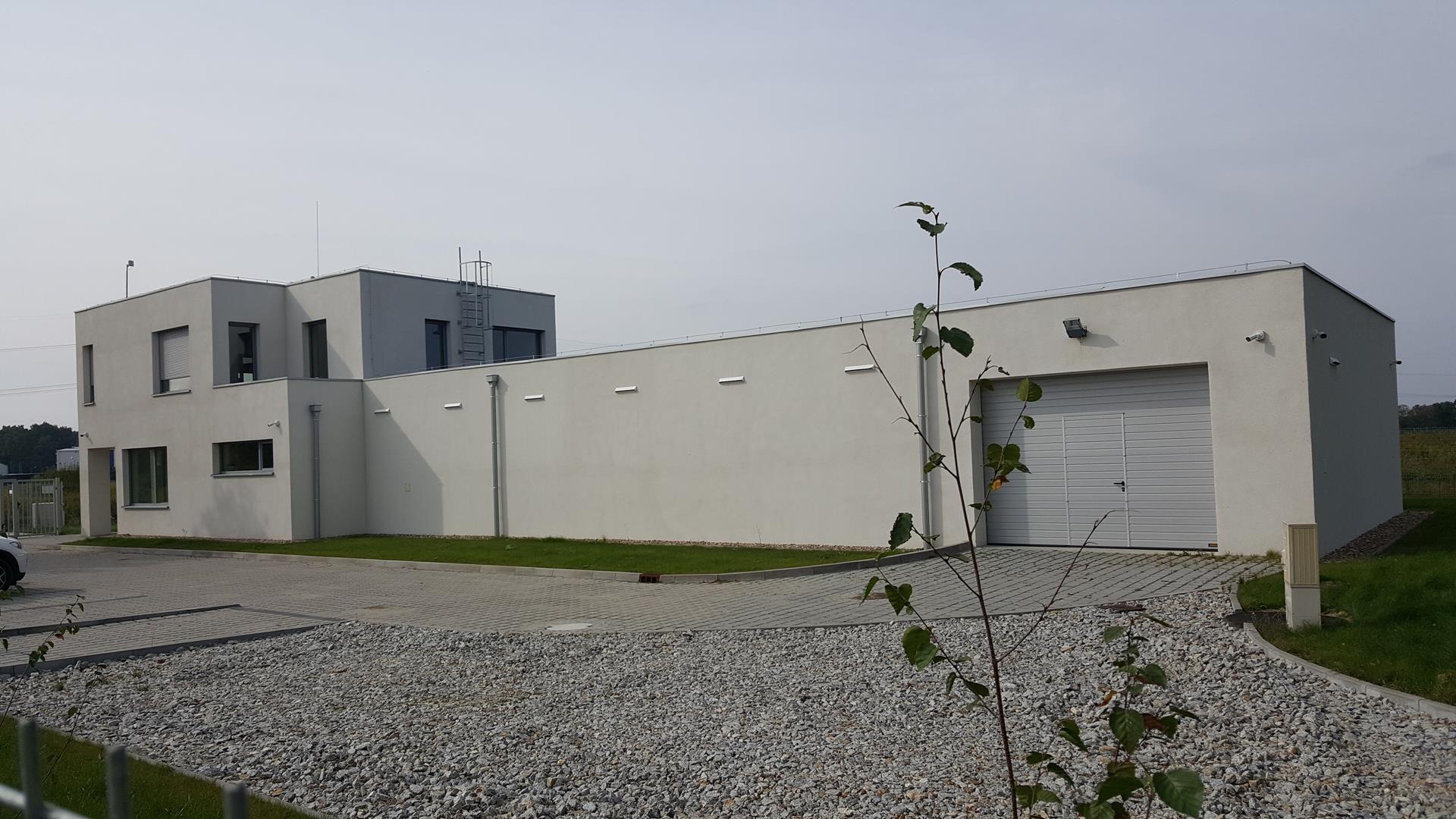 Nowoczesny, dwukondygnacyjny budynek biurowy z szarą elewacją i garażem z bramą segmentową, otoczony utwardzonym terenem z kamienia, widoczne kamery monitoringu.