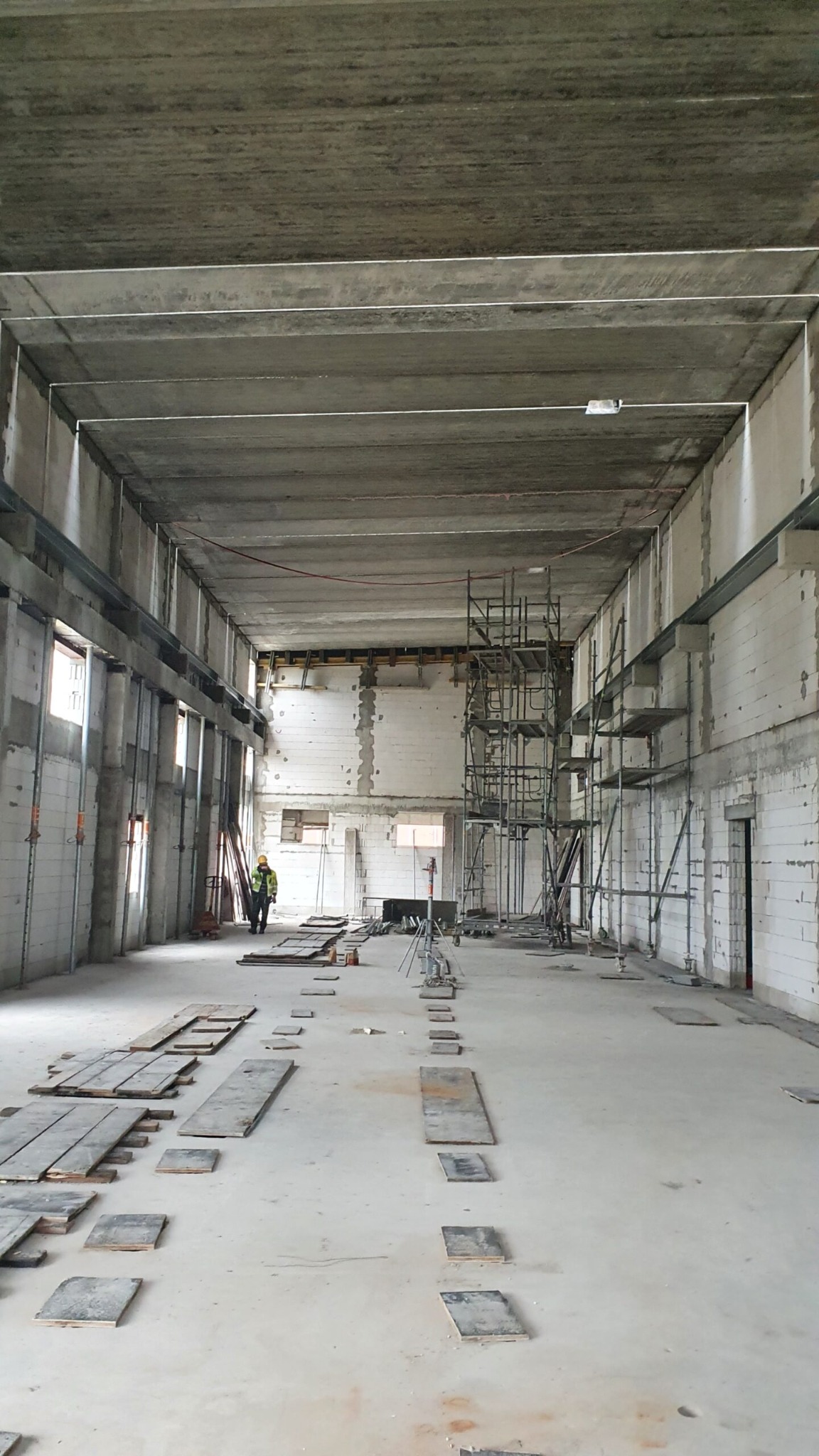 Wnętrze hali w trakcie budowy z widocznymi rusztowaniami, płytami na podłodze i pracownikiem w oddali.