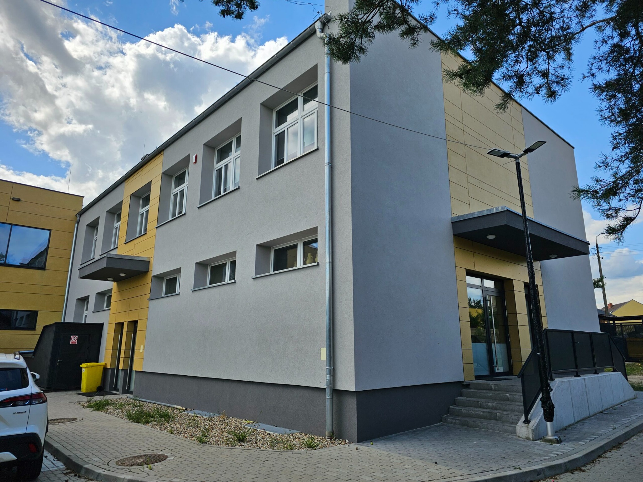Nowoczesny budynek dwukondygnacyjny z szarą elewacją i żółtymi akcentami, widoczny z poziomu chodnika wyłożonego kostką brukową, z elementami zagospodarowania terenu i lampą uliczną.