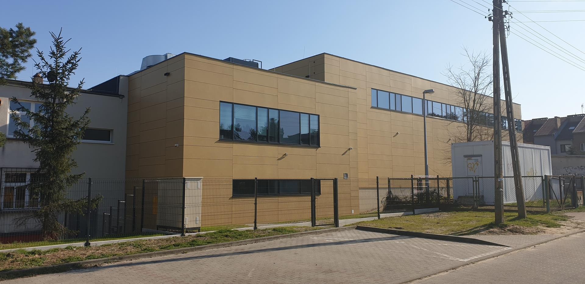 Nowoczesny budynek biurowy z elewacją w odcieniach beżu, widoczny z zewnątrz, z dużymi oknami odbijającymi otoczenie, ogrodzony metalową siatką, z kontenerem i słupami energetycznymi w tle.