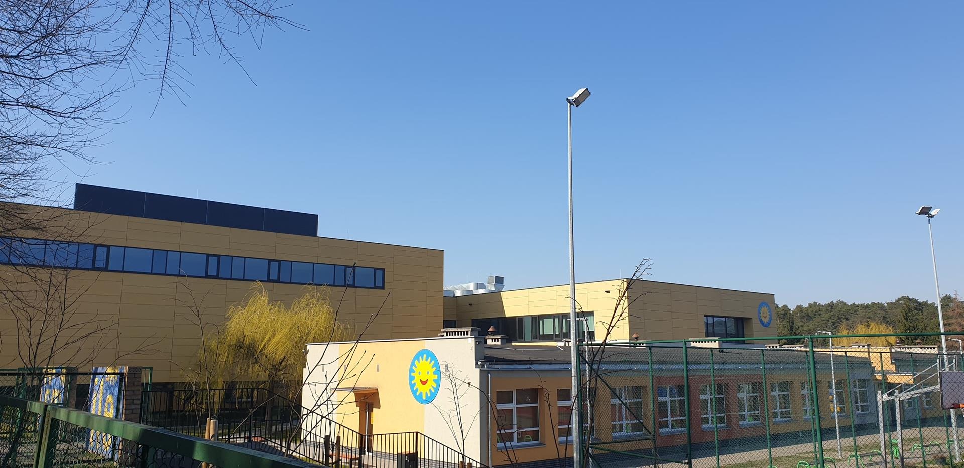 Widok na budynek szkoły z żółtą elewacją i niebieskim logo słońca, otoczony zielonym ogrodzeniem i drzewami bez liści pod błękitnym niebem.