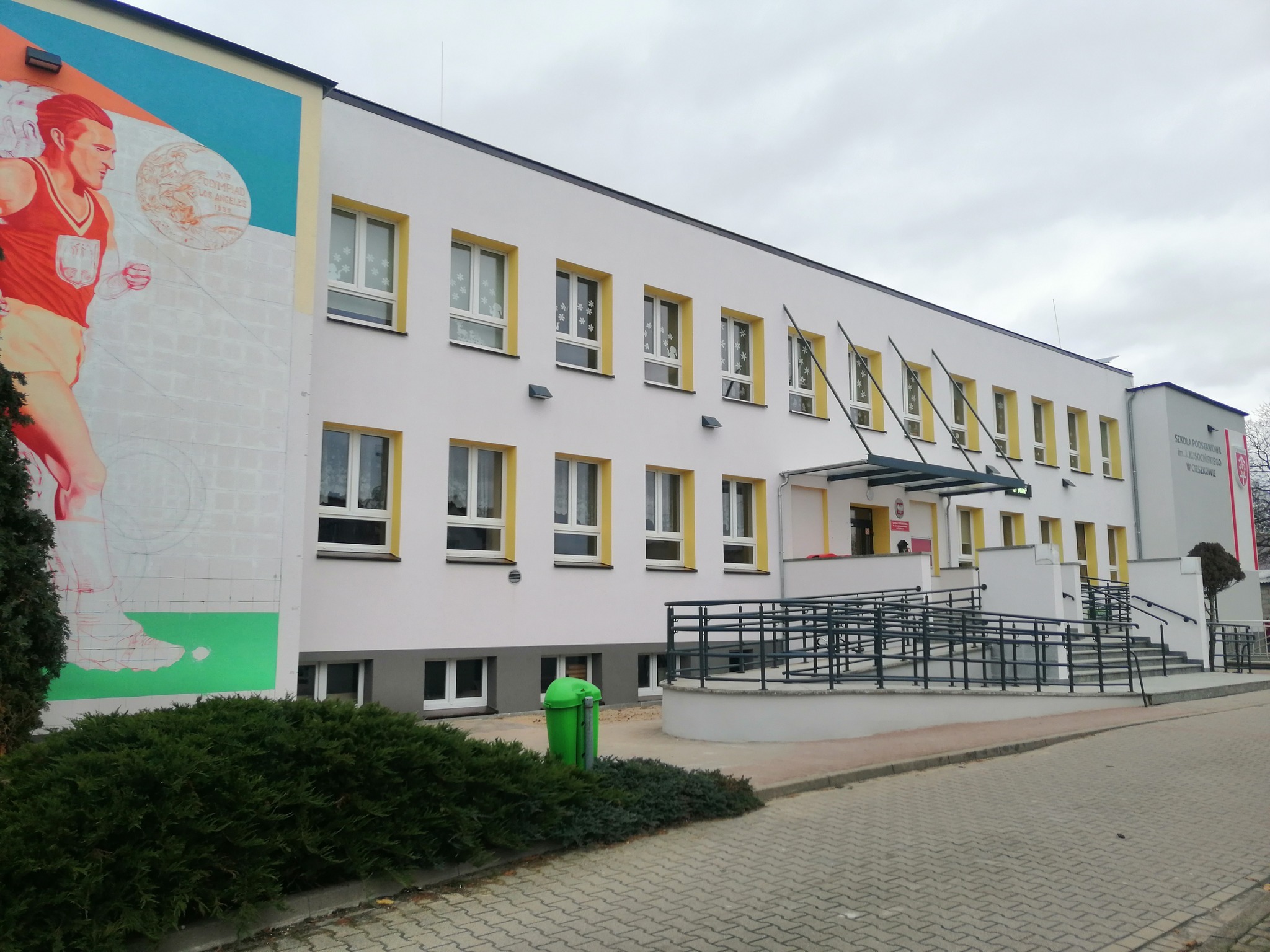 Dwupiętrowy budynek szkoły z białymi ścianami i żółtymi obramowaniami okien, muralem sportowym na bocznej ścianie i rampą dla niepełnosprawnych przy wejściu.