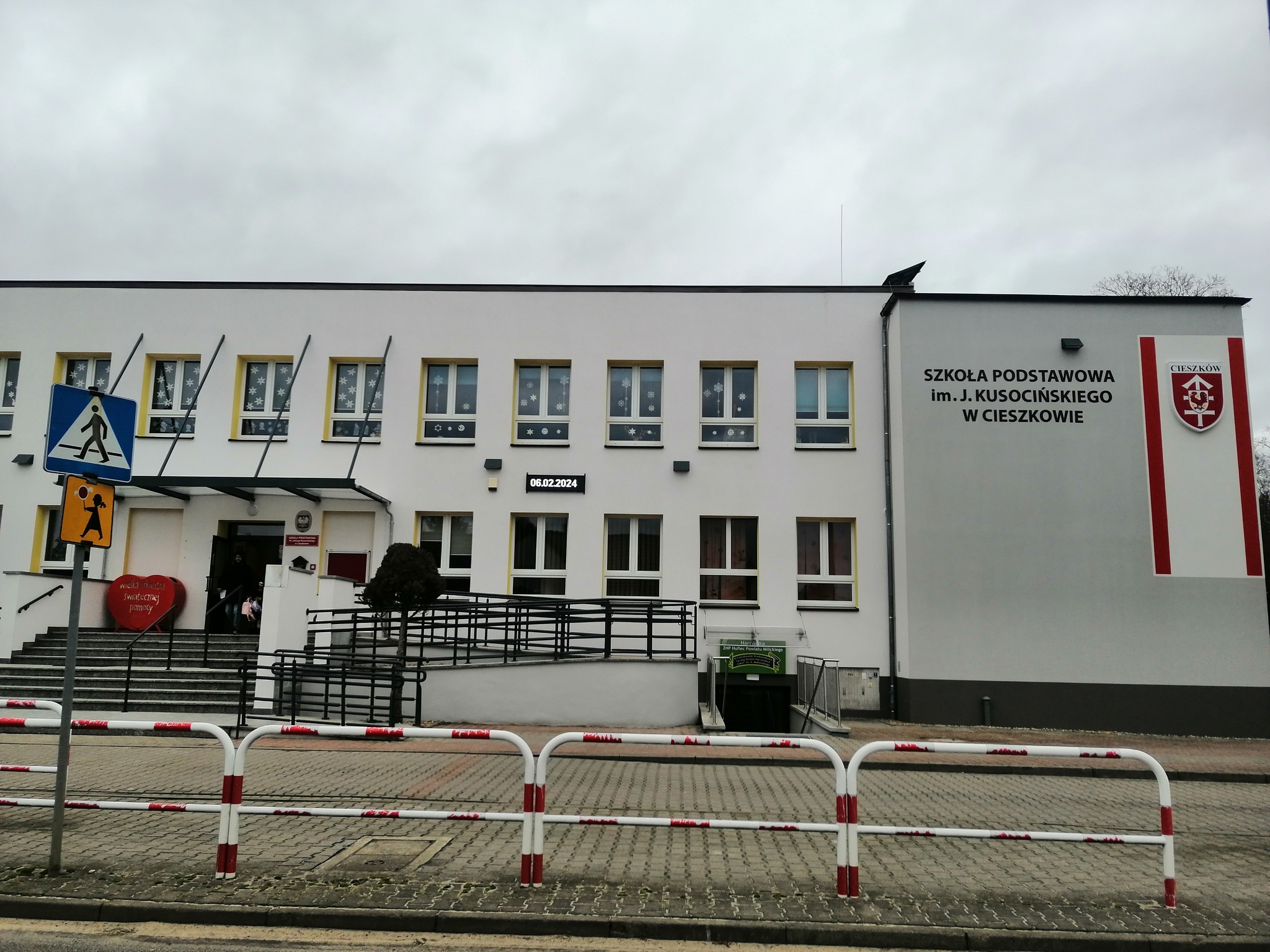Dwukondygnacyjny budynek Szkoły Podstawowej im. J. Kusocińskiego w Cieszkowie z płaskim dachem, białymi ścianami i żółtymi obramowaniami okien, ozdobiony świątecznymi dekoracjami w oknach, widoczny...