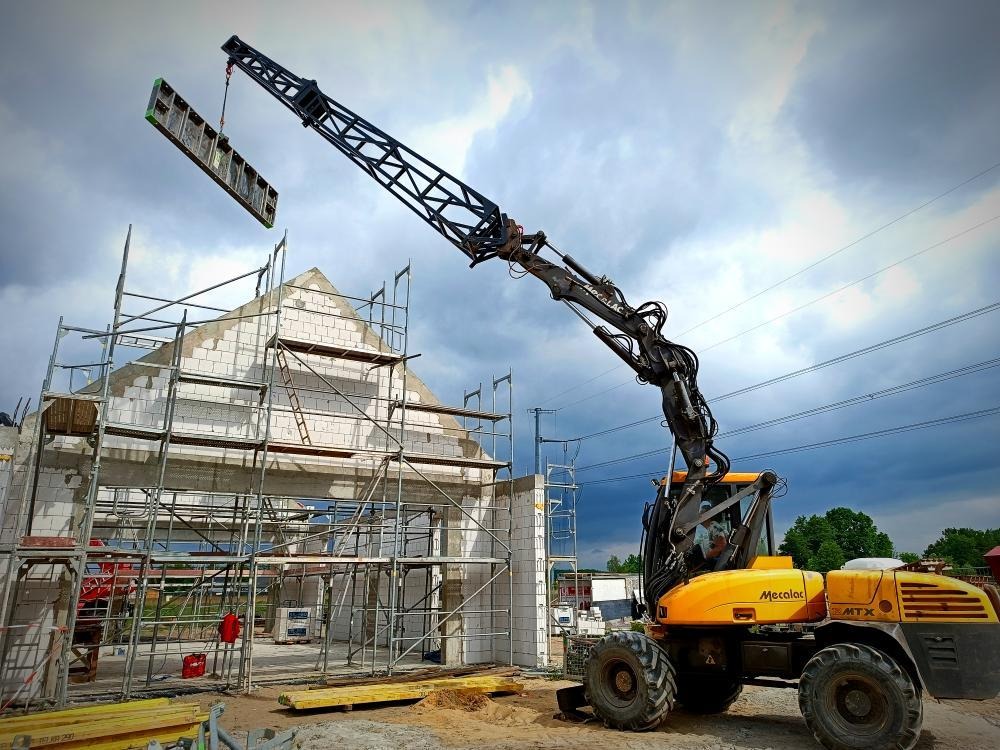 Żółta maszyna budowlana Mecalac podnosi element szalunkowy na budowę domu z białego bloczka, widoczne rusztowania i pochmurne niebo w tle.