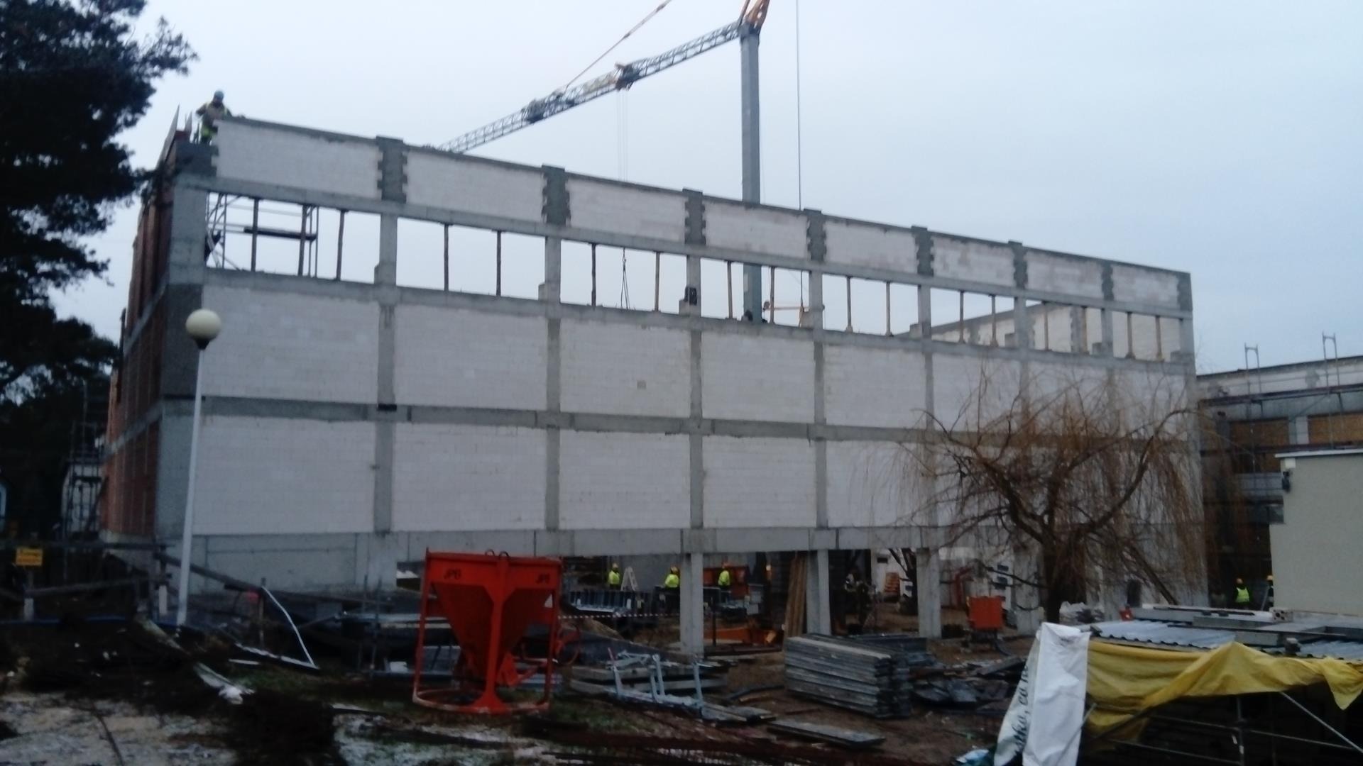 Wznoszenie dwukondygnacyjnej hali z betonu komórkowego, widoczny żuraw budowlany w tle, pracownicy w żółtych kamizelkach na placu budowy oraz czerwony kontener na zaprawę.