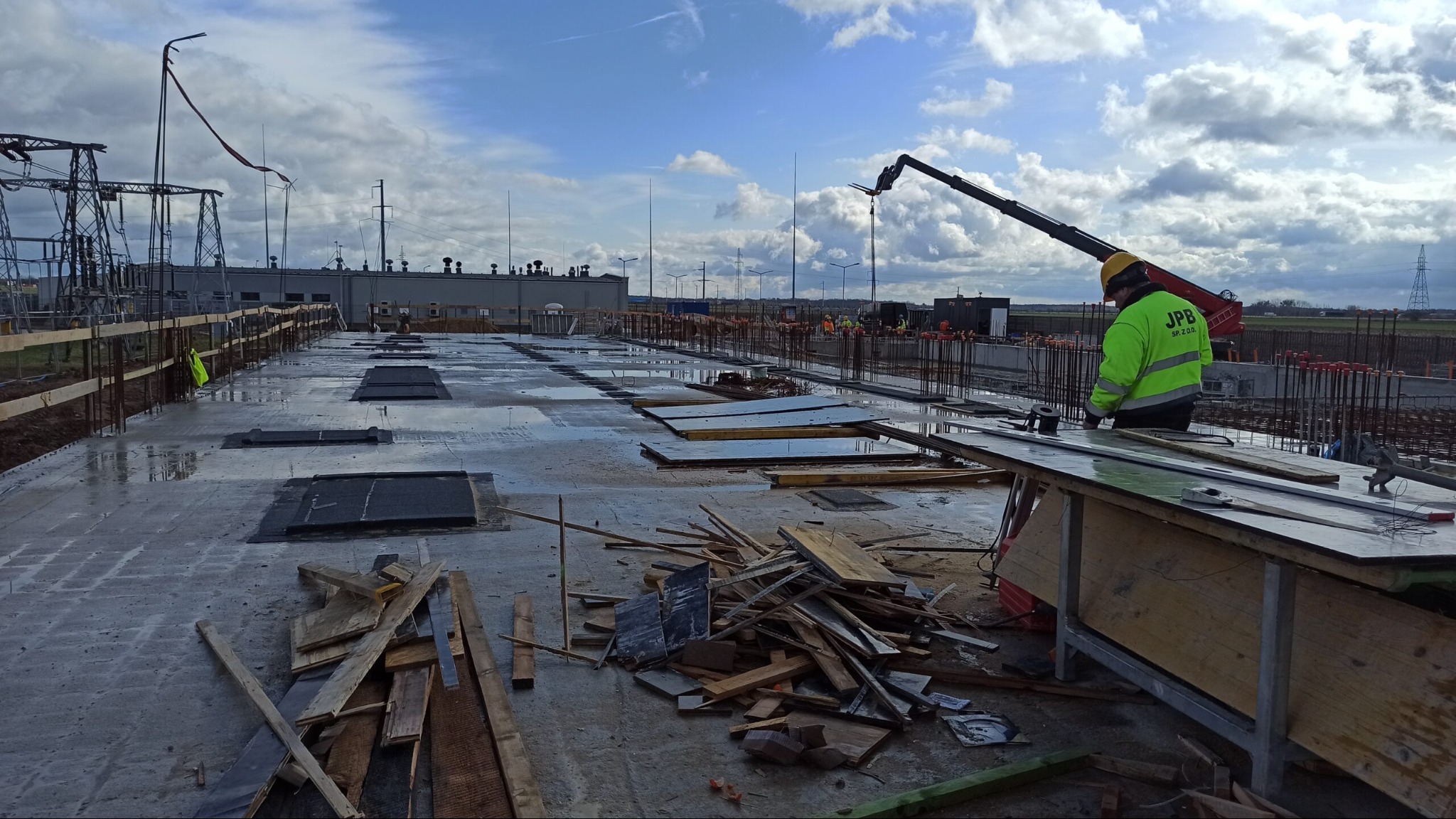 Wylewka betonowa na budowie hali przemysłowej, widoczny pracownik w żółtej kamizelce, zbrojenia, szalunki, żuraw budowlany i elementy infrastruktury energetycznej w tle.