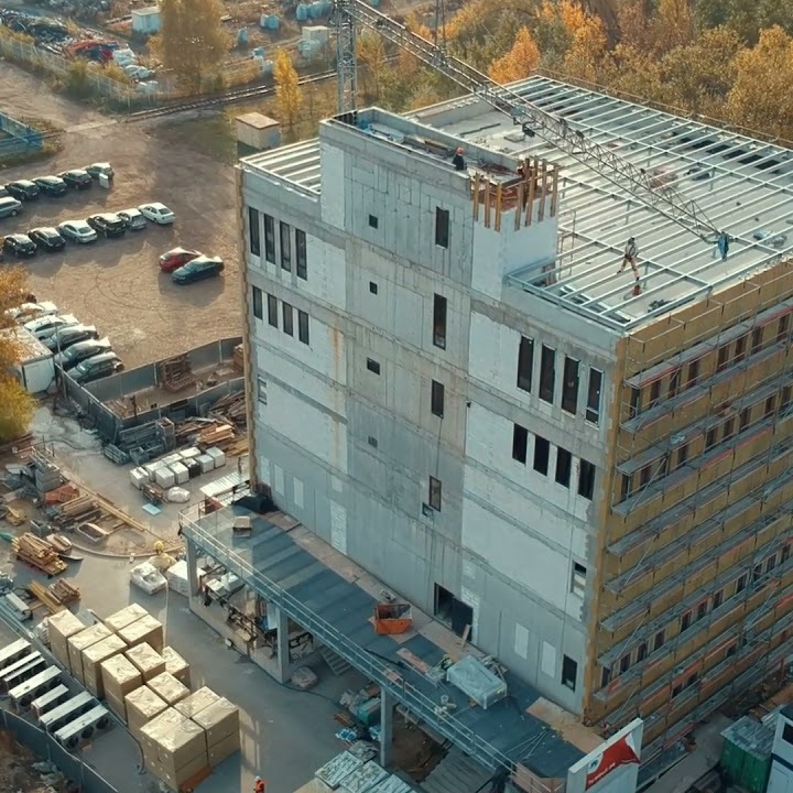 Wysoki budynek w trakcie budowy, widok z góry, z widocznym szkieletem konstrukcji stalowej na dachu i częściowo ocieploną elewacją, na placu budowy zaparkowane samochody i składowane materiały...