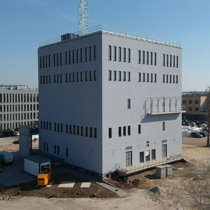 Nowoczesny, sześciopiętrowy budynek biurowy w trakcie wykończenia, z szarą elewacją i rzędami prostokątnych okien, widoczny żółty dźwig i robotnicy przy niwelacji terenu wokół budynku.