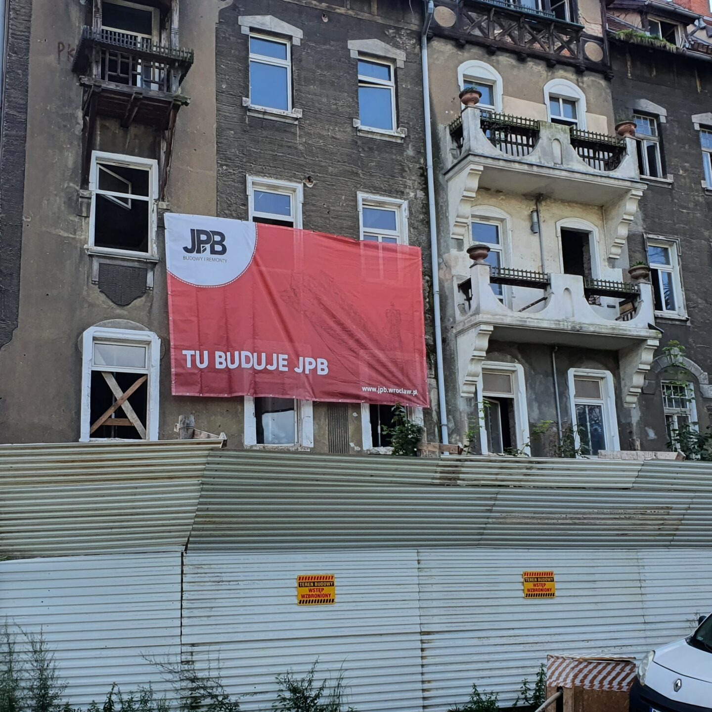 Stara kamienica w trakcie remontu, zabezpieczona blachą falistą i tablicami ostrzegawczymi. Na elewacji budynku wisi duży baner reklamowy firmy budowlanej z logo i napisem 'TU BUDUJE JPB'. Widoczne...