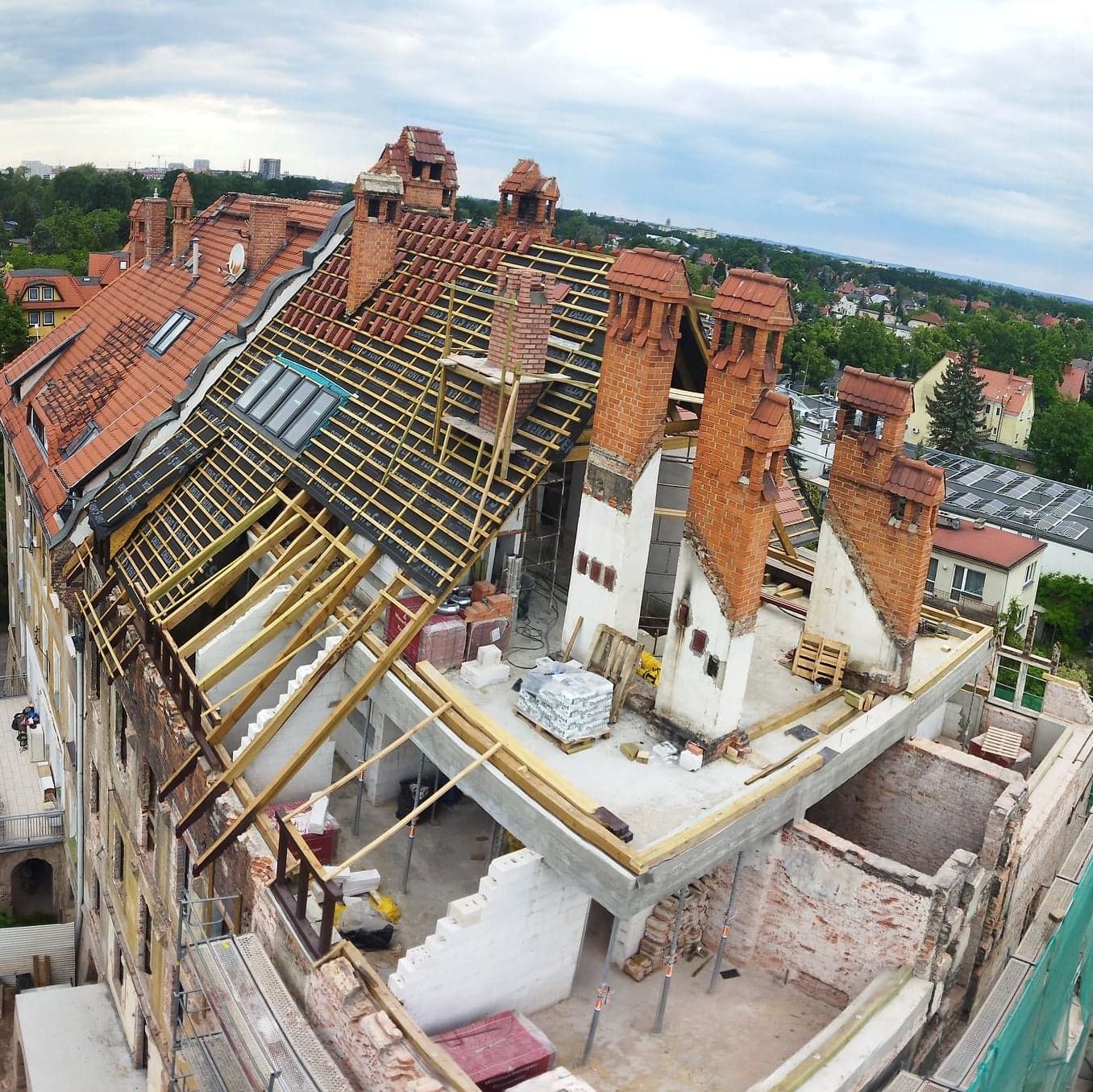 Wysoki kąt widzenia na budynek w trakcie remontu dachu, z widocznymi elementami konstrukcyjnymi, ceglanymi kominami i częściowo ułożoną dachówką, na tle zieleni miejskiej.
