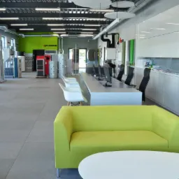 Przestronne wnętrze sklepu z urządzeniami grzewczymi i instalacyjnymi, widoczne ekspozycje kotłów i zbiorników, nowoczesne biuro z zielonym akcentem kolorystycznym na ścianie i sofie.