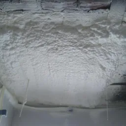 Świeżo nałożona, gęsta, biała izolacja piankowa na suficie z fragmentami odsłoniętej cegły i folią ochronną na ścianie, widoczne niewielkie niedociągnięcia w aplikacji.