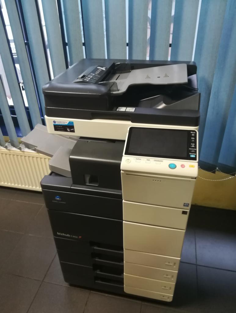 Wielofunkcyjna kserokopiarka biurowa Konica Minolta bizhub C454e stojąca na ciemnej podłodze, z widocznymi zasłonami w tle.