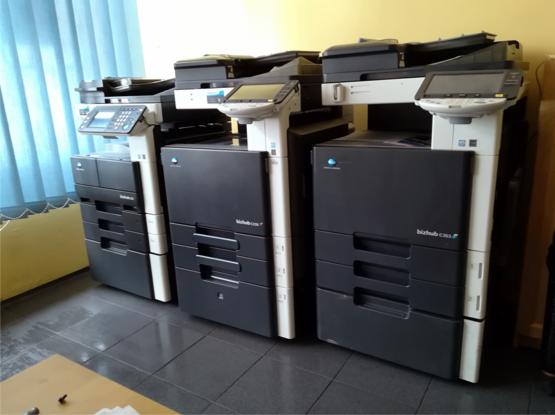 Rząd trzech czarno-białych kserokopiarek Minolta Bizhub C459 z dotykowymi panelami sterowania, ustawionych obok siebie w pomieszczeniu z niebieskimi zasłonami.