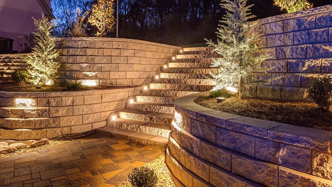 Kaskadowy ogród z kamiennymi schodami i oświetlonymi murkami, ozdobnymi krzewami iglastymi, oświetlenie punktowe na schodach i murach, widok nocny.