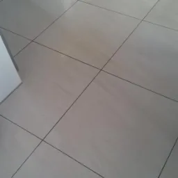 Jasne, kwadratowe płytki podłogowe z ciemnymi fugami, ułożone ukośnie, z widocznym fragmentem białej listwy przypodłogowej i szarej szafki w rogu, oświetlone naturalnym światłem.