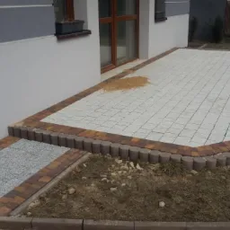 Wykończony taras z jasnoszarej kostki brukowej ułożonej w łuk, obramowany ciemniejszą kostką i palisadą, z fragmentem chodnika z szarego grysu i piaskiem na powierzchni tarasu przy ścianie budynku.