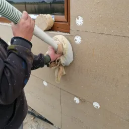 Prace związane z ocieplaniem ściany budynku w Barcinie, widoczne płyty izolacyjne STEICO, mocowane za pomocą łączników, oraz pracownik w trakcie wdmuchiwania izolacji w szczelinę.