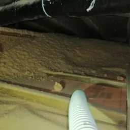 Aplikacja izolacji natryskowej w trudno dostępnej przestrzeni poddasza, widoczne rury instalacyjne i drewniana konstrukcja dachu.