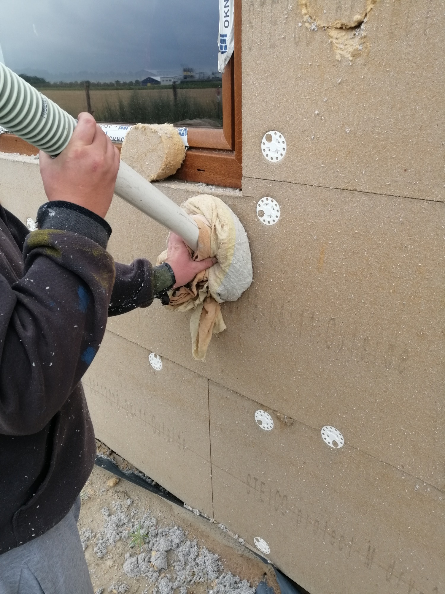 Prace związane z ocieplaniem ściany budynku w Barcinie, widoczne płyty izolacyjne STEICO, mocowane za pomocą łączników, oraz pracownik w trakcie wdmuchiwania izolacji w szczelinę.