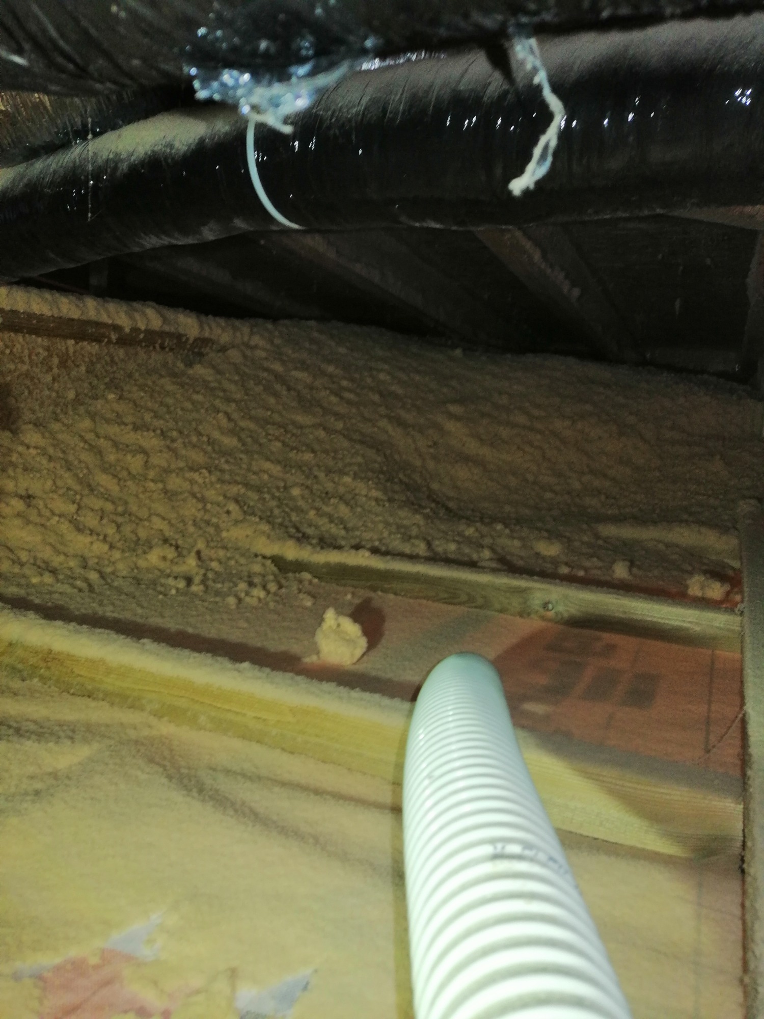 Aplikacja izolacji natryskowej w trudno dostępnej przestrzeni poddasza, widoczne rury instalacyjne i drewniana konstrukcja dachu.