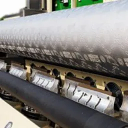 Duży, srebrny walec z wytłoczonym wzorem i napisami umieszczony w maszynie do pikowania ultradźwiękowego tkanin, widoczne elementy sterujące maszyny i wentylatory chłodzące.