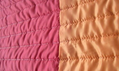 Ultrasonically quilted fabric sample in two colors: coral pink and apricot orange, showing horizontal stitching lines and raised, padded texture.