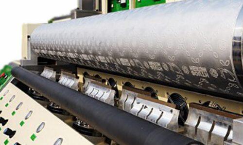 Duży, srebrny walec z wytłoczonym wzorem i napisami umieszczony w maszynie do pikowania ultradźwiękowego tkanin, widoczne elementy sterujące maszyny i wentylatory chłodzące.