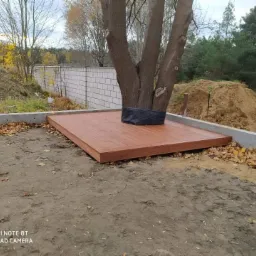 Niski taras z brązowych desek otaczający pień drzewa, zabezpieczony czarną siatką, na tle muru z białej cegły i hałdy ziemi.