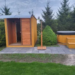 Drewniana sauna z otwartymi drzwiami, zewnętrzny prysznic z deszczownicą i drewniana balia z pokrywą na tle zielonych krzewów i drzew iglastych, ułożone na szarych płytach chodnikowych.