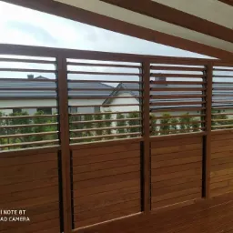 Drewniana pergola z lamelami i drewnianym tarasem, widok na dom z ogrodem w tle, pochmurne niebo.