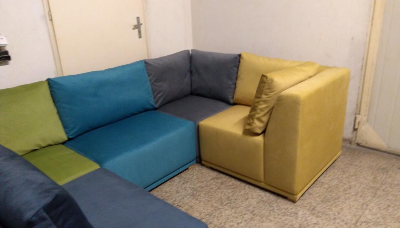 Modułowa sofa narożna z tapicerką w odcieniach zieleni, błękitu, szarości i żółci, ustawiona na tle ściany w pomieszczeniu.