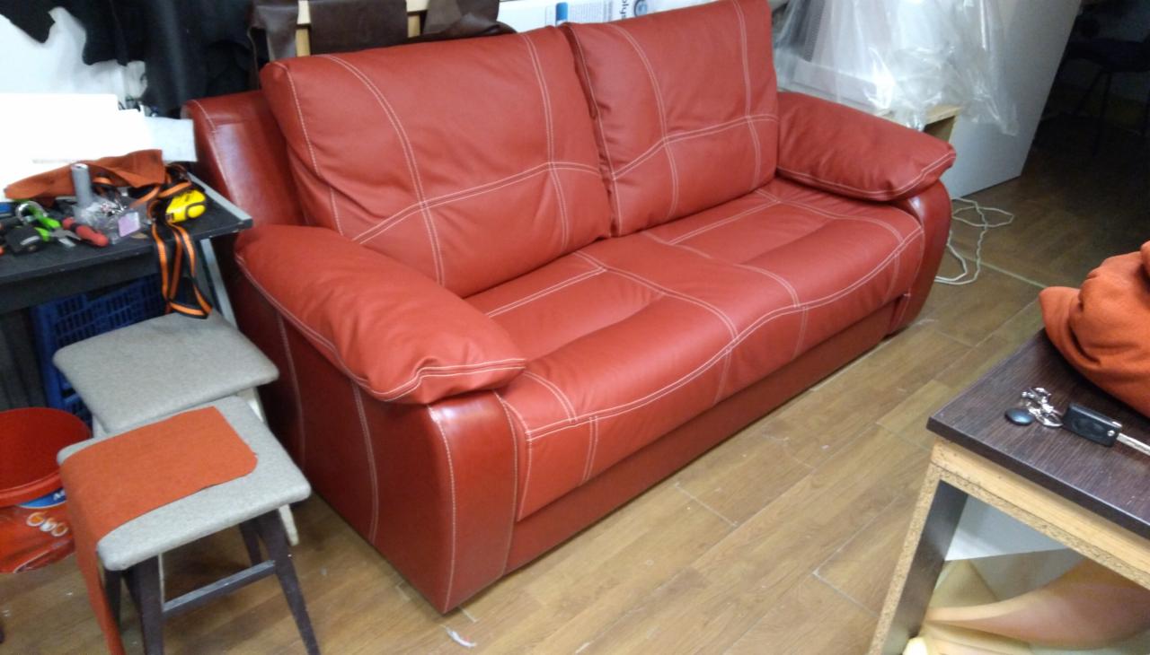 Czerwona skórzana sofa z białymi przeszyciami, widoczna w warsztacie obok stołu z narzędziami i krzesłami w trakcie renowacji.