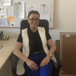 Kobieta w okularach siedzi na krześle biurowym, w tle tablica z notatkami i kalendarzem. Ubrana w czarną bluzkę i białą kamizelkę. Na biurku widoczne przybory.