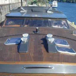 Drewniany pokład jachtu w Szczecinie, widok z przodu z elementami wyposażenia i kabiną, w tle budynek portowy i woda.