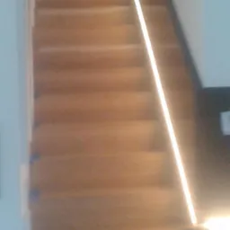 Widok z dołu na drewniane schody z oświetleniem LED wzdłuż boku, prowadzące do góry, z białymi ścianami po bokach.