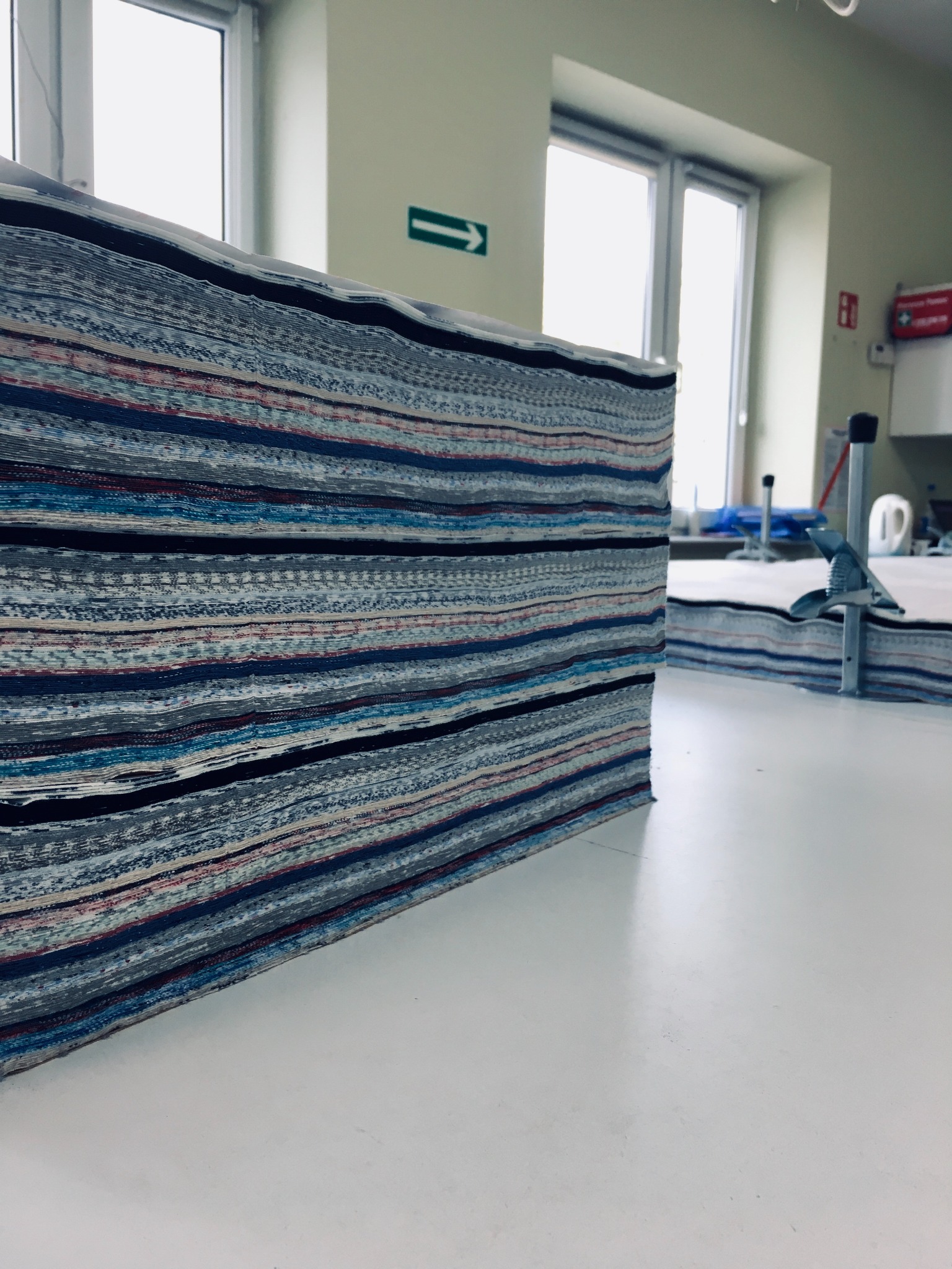 Stos pociętych warstw tkaniny w paski, przygotowanych do dalszej obróbki, leżący na białym stole w jasnym pomieszczeniu. W tle widoczne okno i wyposażenie pracowni.
