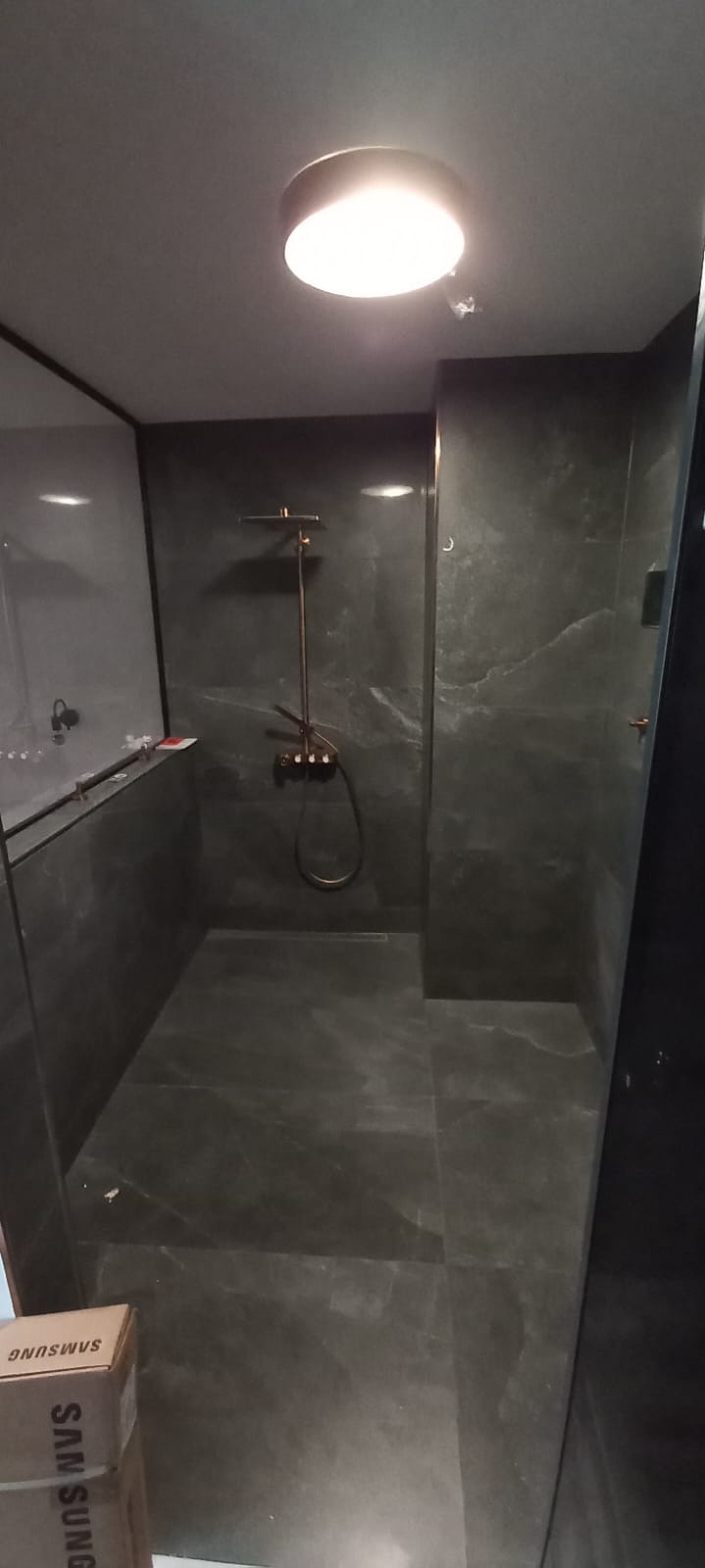 Nowoczesna kabina prysznicowa z ciemnymi płytkami imitującymi marmur, deszczownicą w odcieniach miedzi i subtelnym oświetleniem sufitowym. Widoczny fragment kartonu.