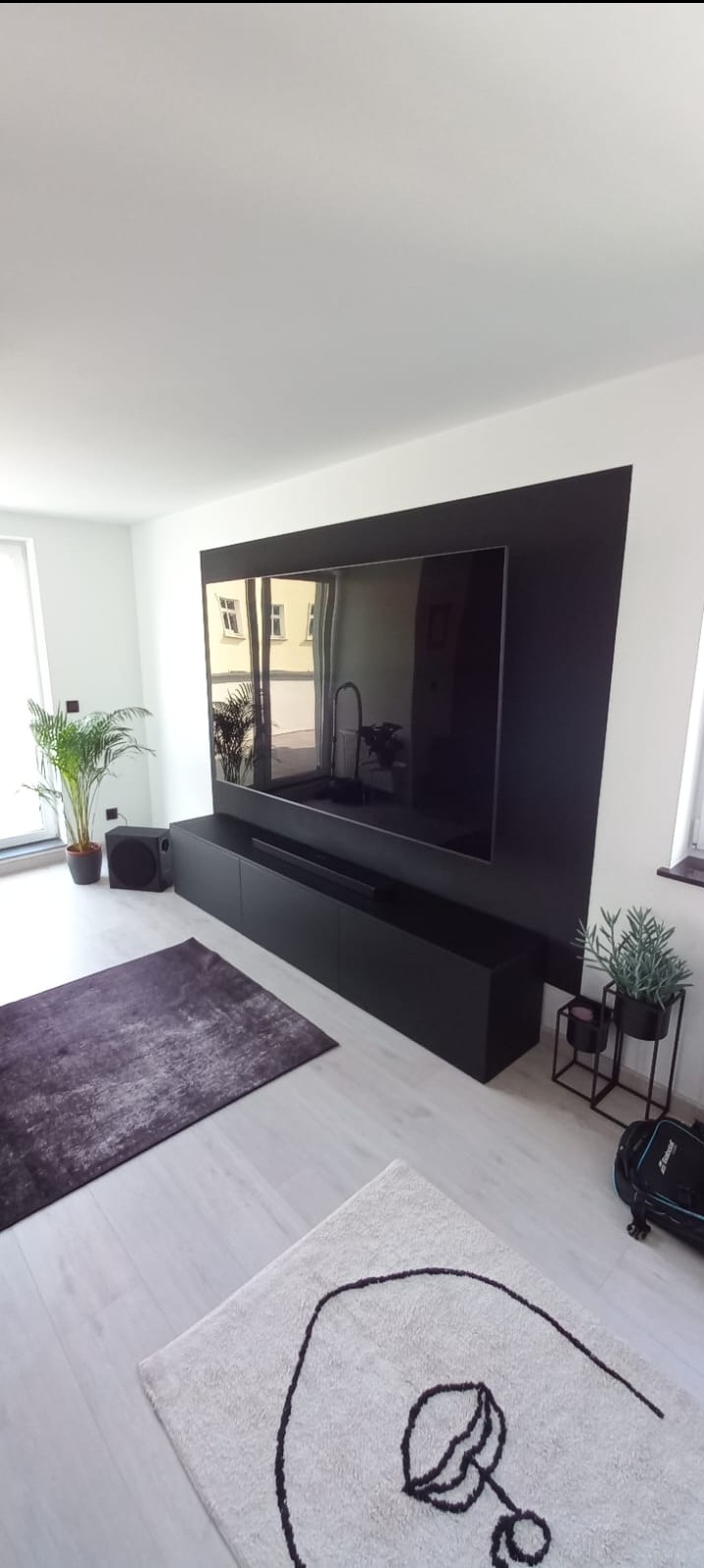 Nowoczesny salon z czarną ścianą TV, minimalistyczną szafką RTV, dywanami i roślinami. W lustrzanej powierzchni telewizora odbija się wnętrze mieszkania.